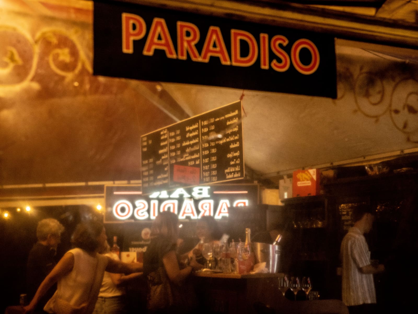 Die Bar Paradiso bei Nacht: Gäste am Tresen, beleuchtete Getränkekarte, warmes Licht